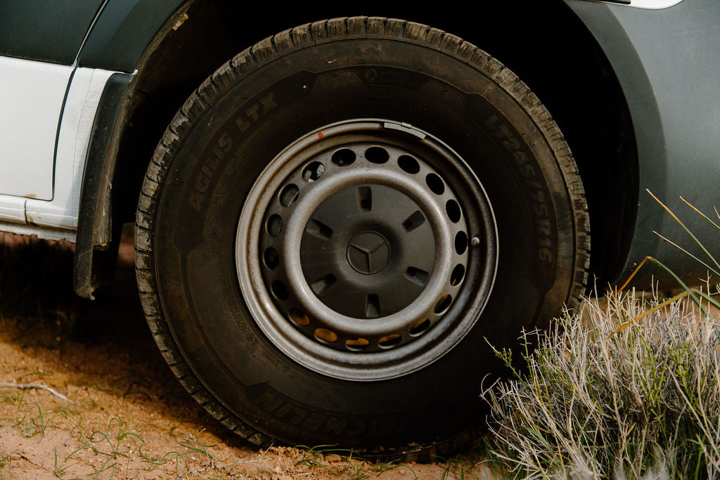 Close-up of an RV rental van's tire and wheel on a dirt path, a Mercedes logo visible on the hubcap.