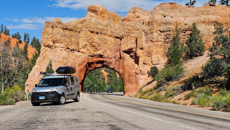Silver RV rental van with roof box parked by a scenic red rock tunnel. A perfect stop on a sunny national park road trip.|Experience van life in this RV rental! Cozy bed — photo 1