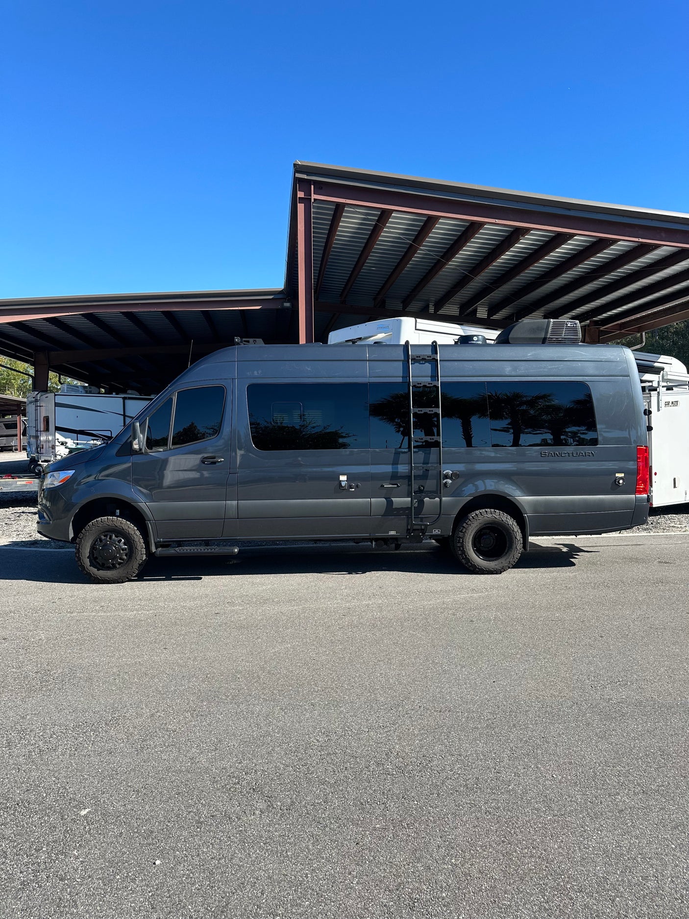 A dark gray Sprinter-style camper van, labeled "Sanctuary", parked under covered storage. Perfect for rv rental trips.