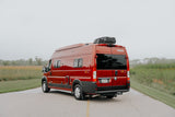 A bright red Winnebago Solis camper van, perfect for RV rental, rests on a road, flanked by green fields under an overcast sky.