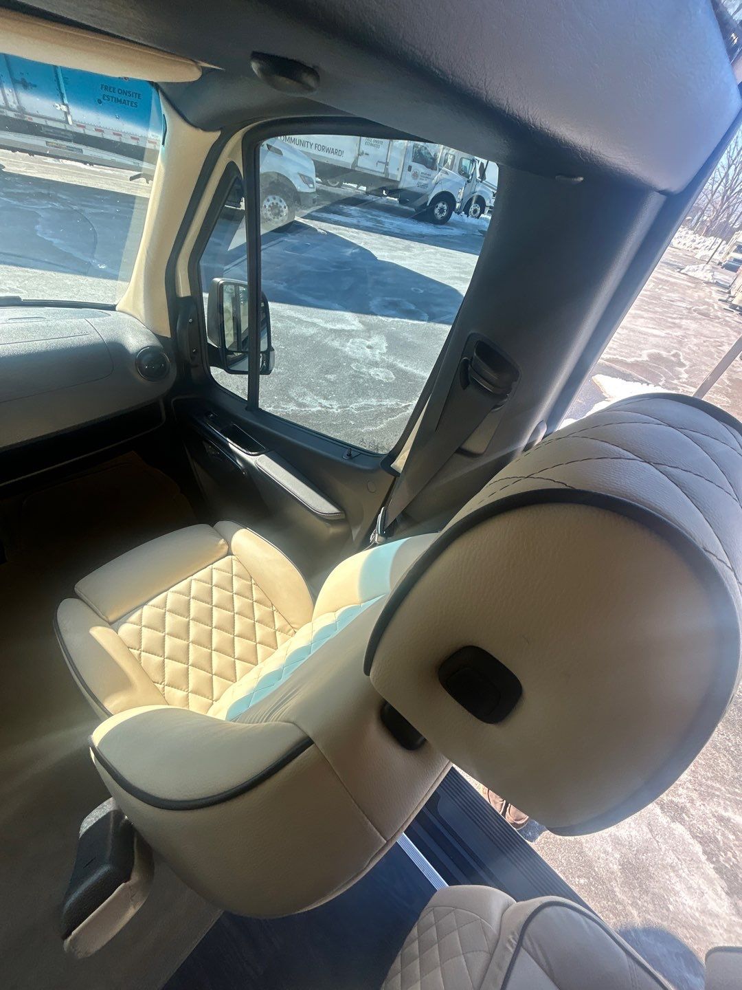 Inside a bright **rv rental**: view of cream quilted captain's seats, looking out at trucks on a sunny lot.