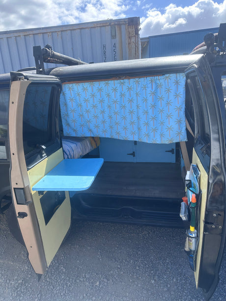 This rv rental features a custom camper van interior: blue palm tree curtain, bed, and a handy fold-down table.