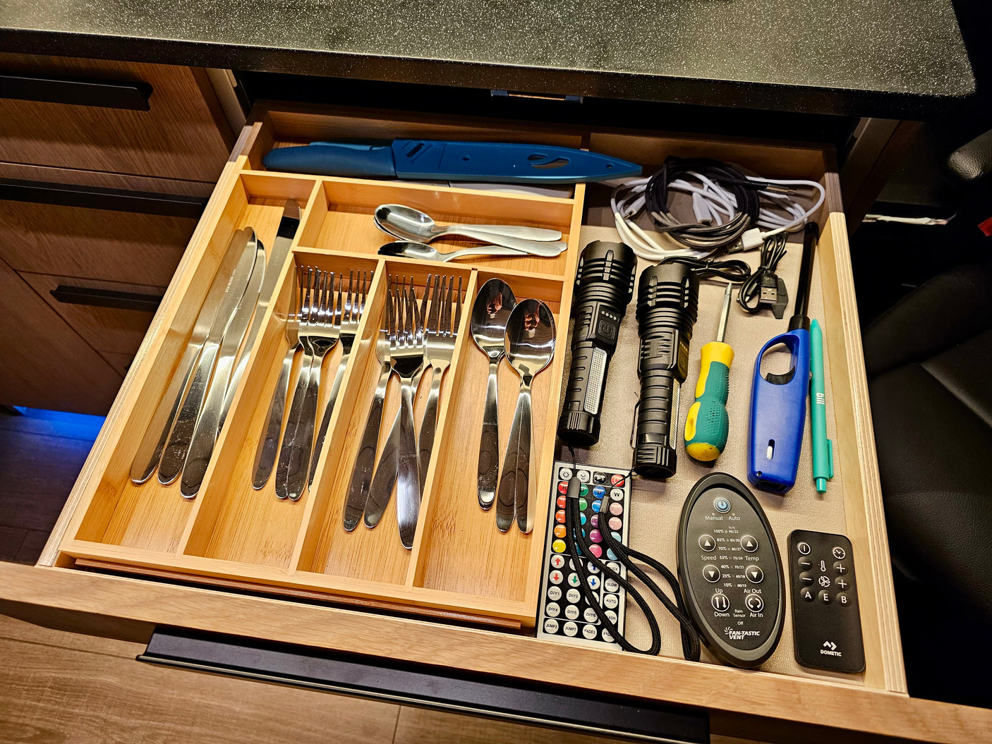 Organized RV rental kitchen drawer: utensils, knife, charging cables, flashlights, screwdriver, and remotes for vent/lights.