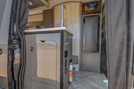 Compact rv rental kitchen with a sink, microwave, fridge, and fire extinguisher in a modern van interior.