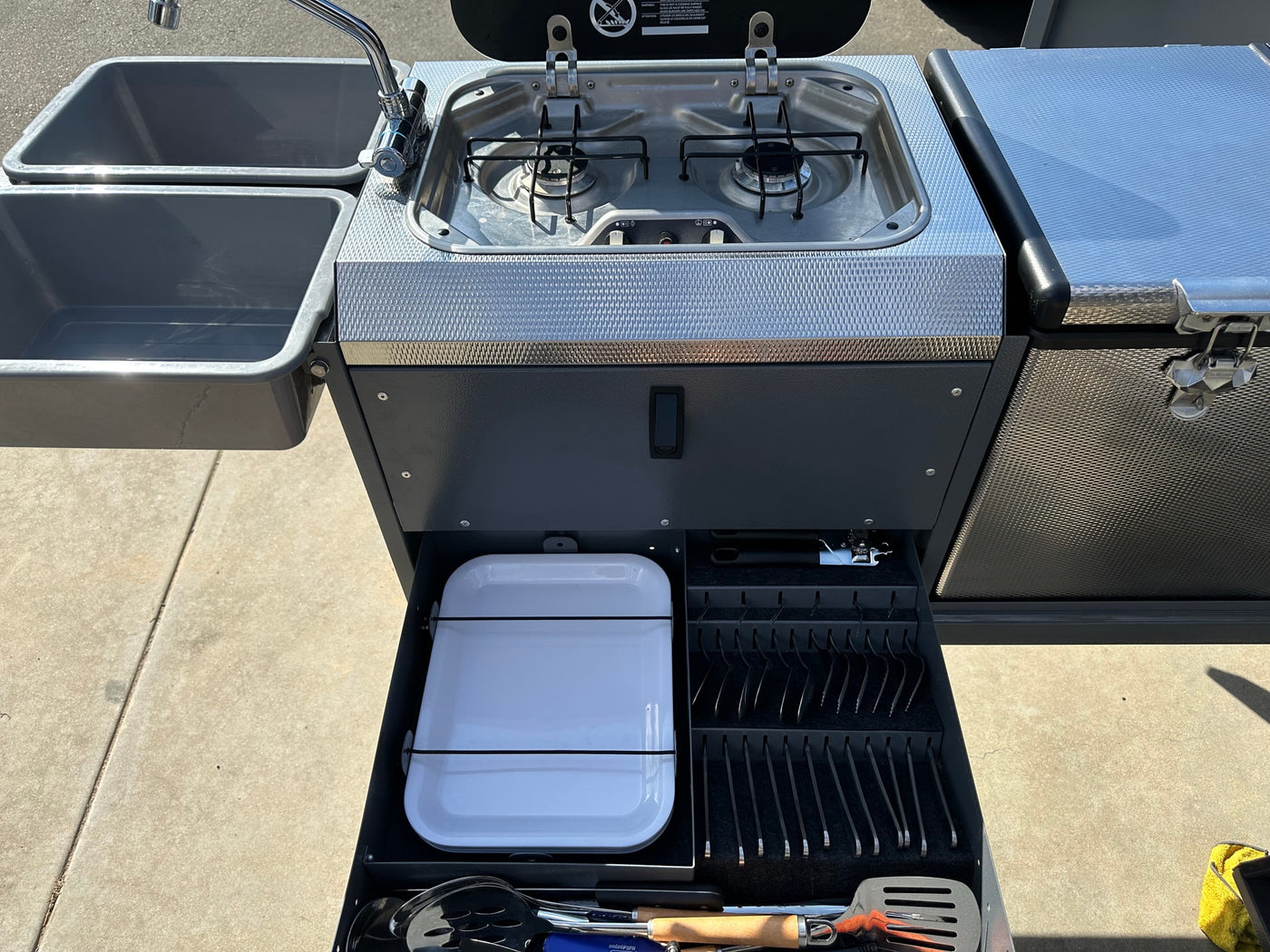 This RV rental features a compact outdoor kitchen with dual sinks, a two-burner stove, and a fully stocked utensil drawer. — photo 14
