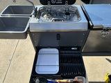 This RV rental features a compact outdoor kitchen with dual sinks, a two-burner stove, and a fully stocked utensil drawer.