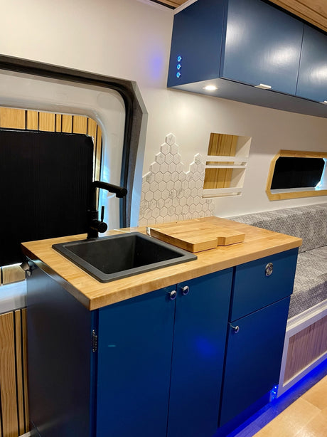 Stylish RV rental interior: blue cabinets, wood counter, black sink, modern backsplash, and cozy seating area.