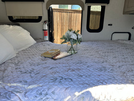 A comfortable bed with a quilted spread, white pillows, books, and flowers inside an RV rental. A fire extinguisher is mounted nearby.