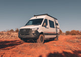 An adventure-ready Storyteller Overland Mercedes Sprinter van, perfect for rv rental, parked on red dirt in a sunny, dry landscape.