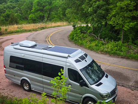 Silver Mercedes Sprinter rv rental with solar panels, parked on a gravel shoulder next to a winding road in a lush forest.