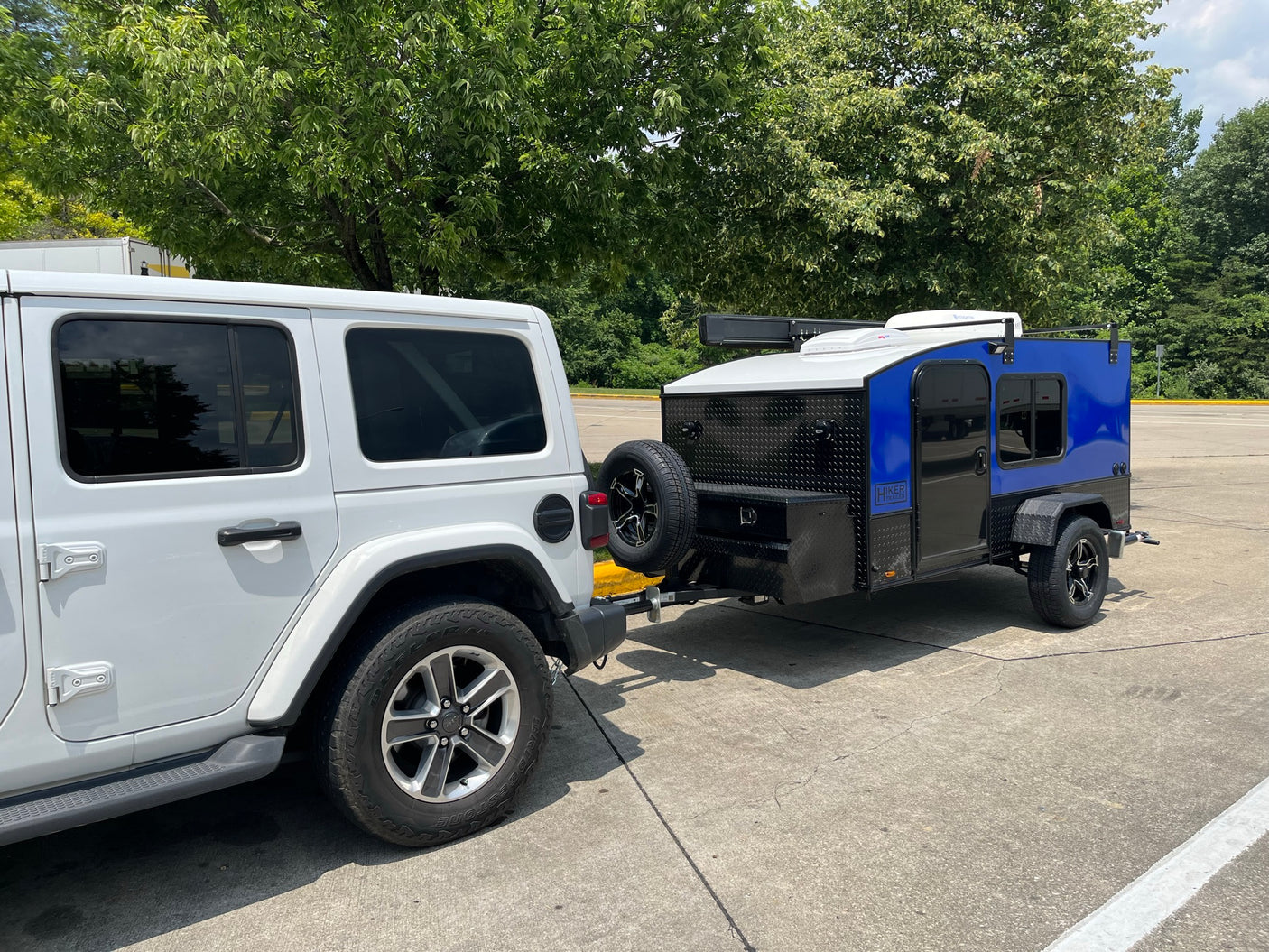A white Jeep Wrangler tows a compact, blue and black Hiker Trailer, perfect for your next RV rental adventure.