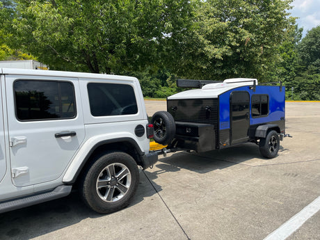 A white Jeep Wrangler tows a compact, blue and black Hiker Trailer, perfect for your next RV rental adventure.