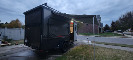 Dark grey RV rental camper trailer with an extended awning, parked on a driveway. Ready for outdoor travel adventures.
