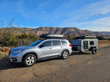 Silver SUV towing a grey teardrop camper with rooftop gear in a mountain landscape, great for an RV rental trip.