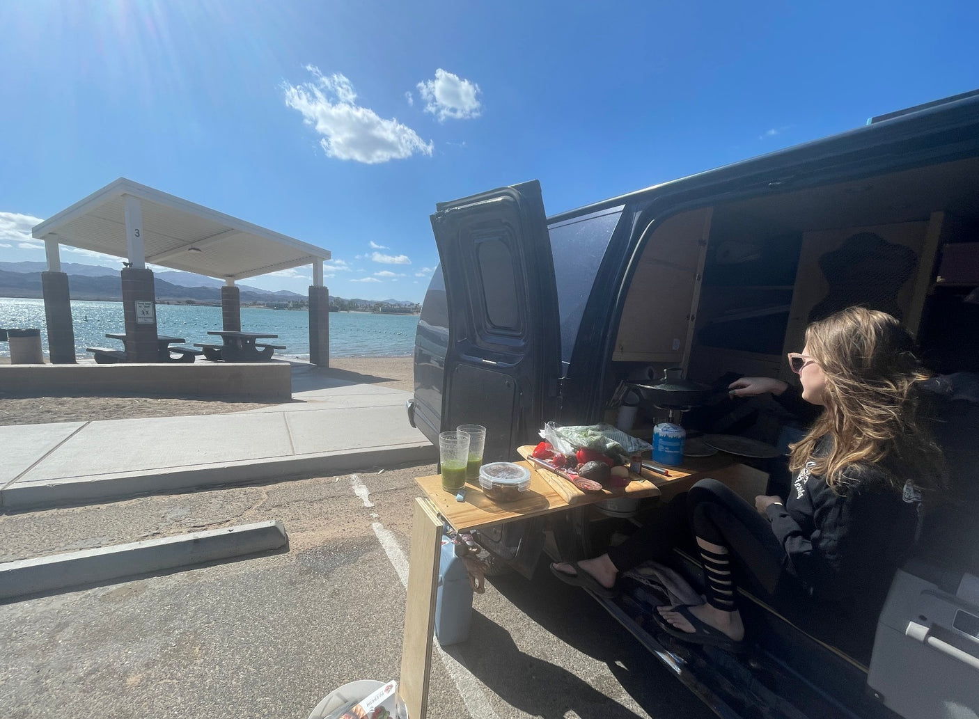 Cooking fresh meals from an **rv rental** van by a sunny lake. A woman relaxes at her mobile camp, enjoying the view.