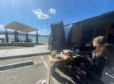 Cooking fresh meals from an **rv rental** van by a sunny lake. A woman relaxes at her mobile camp, enjoying the view.