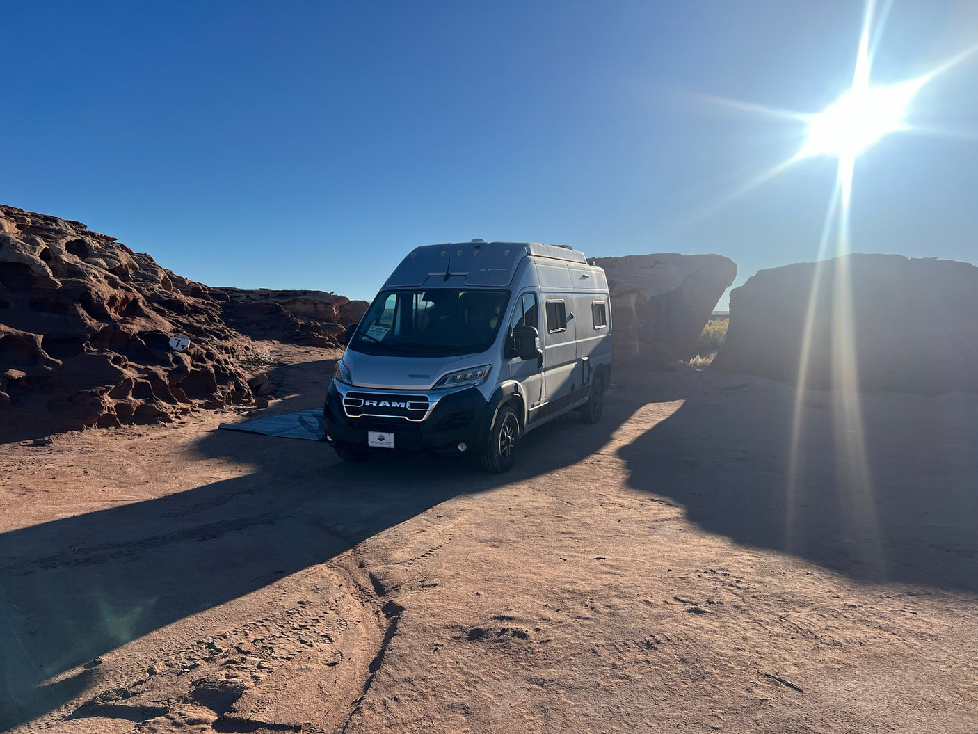 RV rental: A silver camper van parked on red dirt, with sunny red rock formations and long shadows, ideal for a desert adventure.