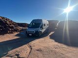 RV rental: A silver camper van parked on red dirt, with sunny red rock formations and long shadows, ideal for a desert adventure.