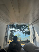 Inside an RV rental, a person is relaxing on the bed, enjoying a beautiful view of a lake through the open back doors.