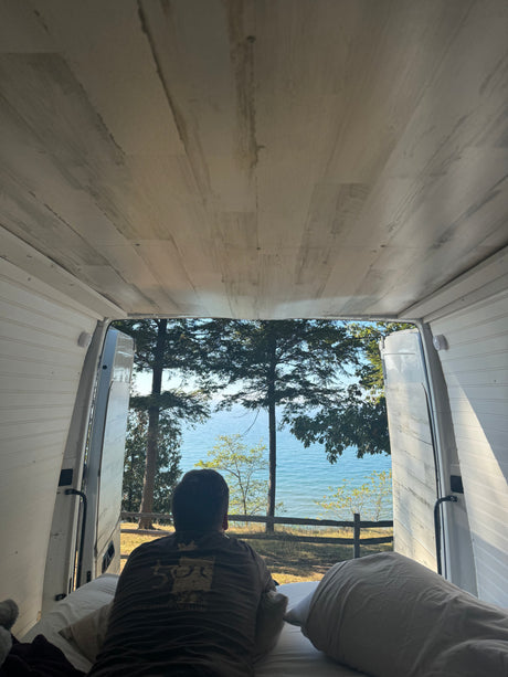 Inside an RV rental, a person is relaxing on the bed, enjoying a beautiful view of a lake through the open back doors.