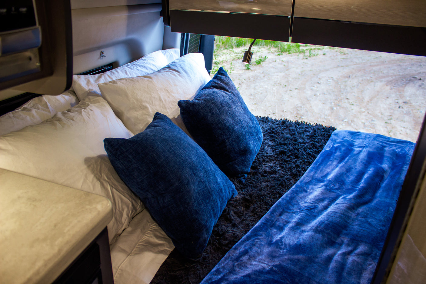 Cozy RV rental bedroom with white sheets, plush blue pillows & blanket, plus an outdoor view.