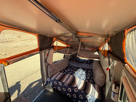 Cozy rv rental interior with a bed, two bunk beds, and windows showing outdoor landscape. A fire extinguisher is mounted inside.