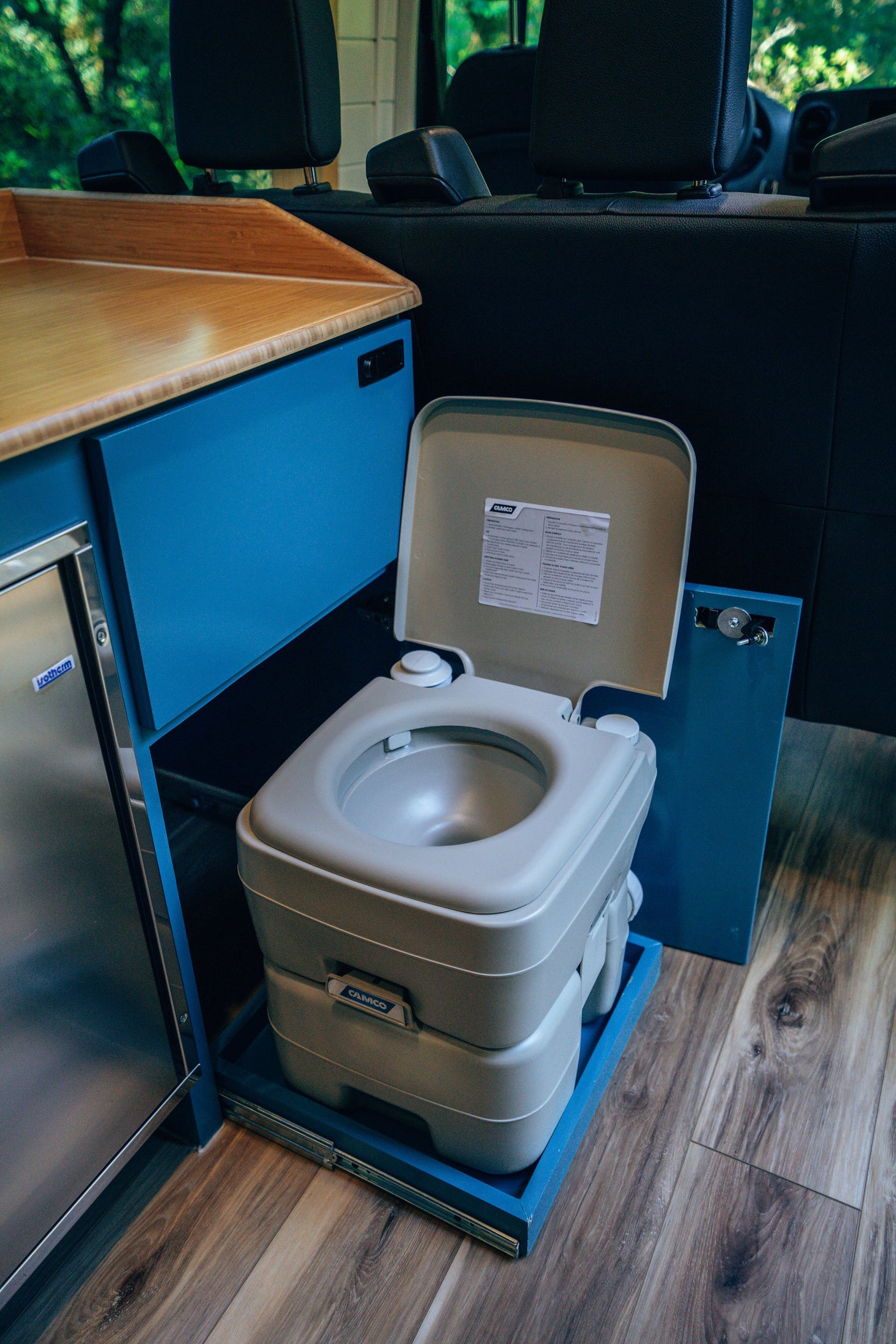 RV rental interior featuring a portable Camco toilet on a pull-out tray, alongside a sleek wood counter, blue cabinetry, and an Isotherm fridge.