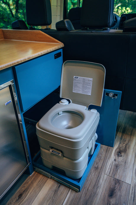 RV rental interior featuring a portable Camco toilet on a pull-out tray, alongside a sleek wood counter, blue cabinetry, and an Isotherm fridge.