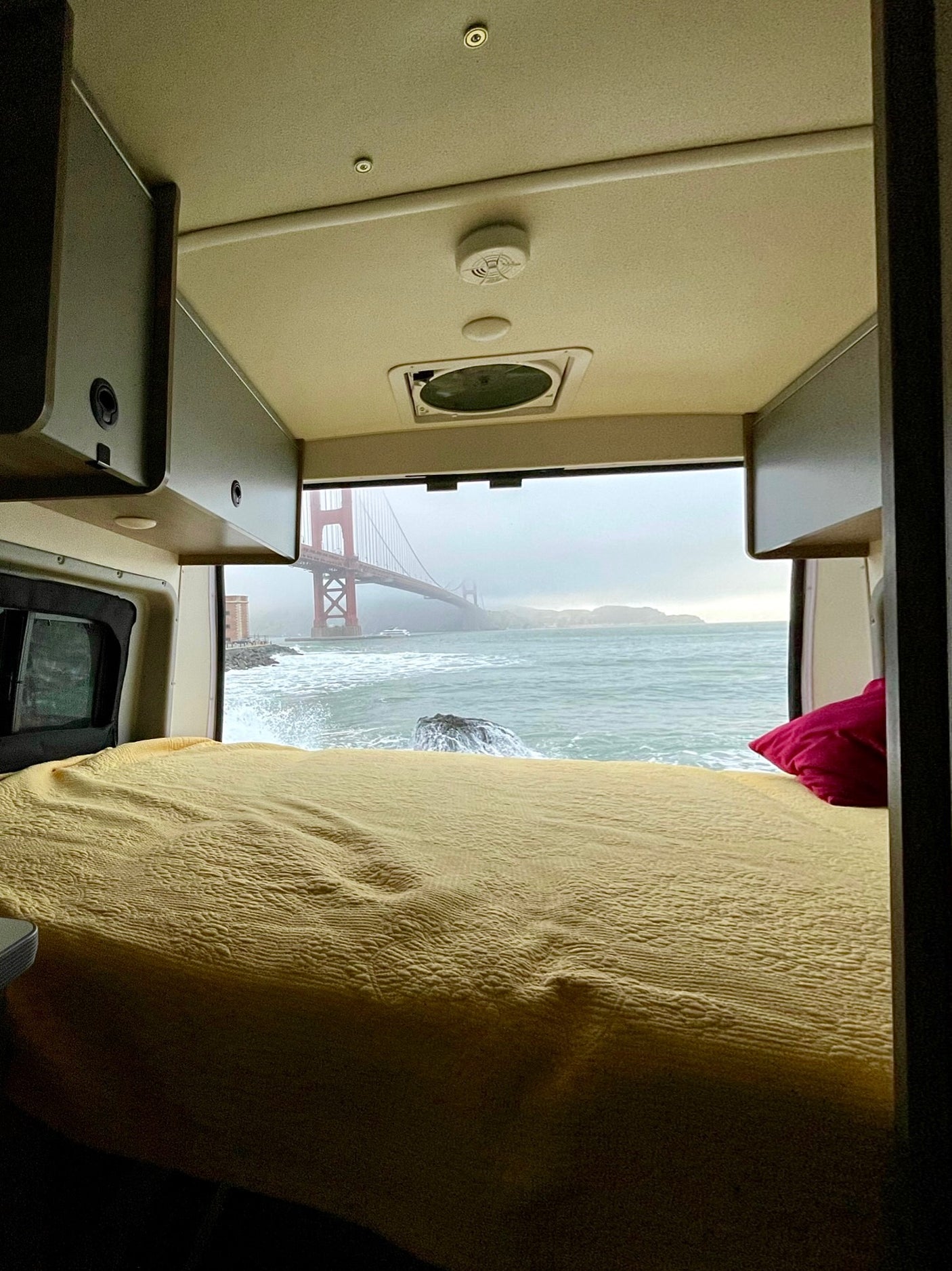 An RV rental bed with a yellow quilt faces a window overlooking the foggy Golden Gate Bridge and crashing ocean waves. Ideal RV travel.