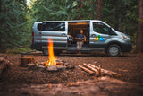RV rental campervan parked in a lush forest. A man plays ukulele inside the van while a campfire crackles nearby.