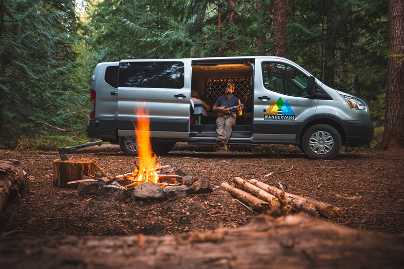 RV rental campervan parked in a lush forest. A man plays ukulele inside the van while a campfire crackles nearby.|This smiling couple and their dog are enjoying their RV rental! Posing with a white converted camper van and open doors.|A woman sits in her open RV rental camper van — photo 1