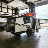 White & black Cricket travel trailer with orange pop-top roof, ideal for rv rental, in a garage bay after a wash.
