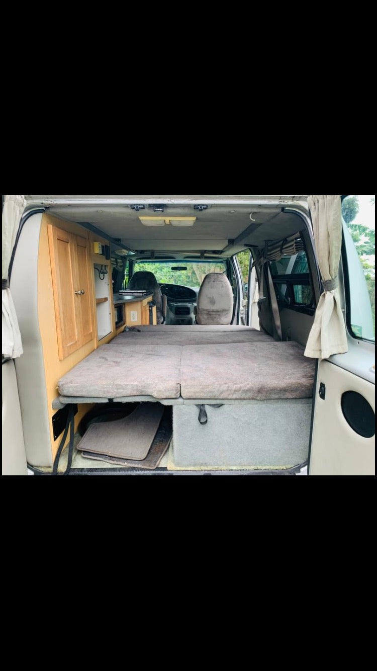 RV rental: Interior of a comfortable camper van with a full bed, light wood cabinetry, & front seats visible from the back. — photo 9