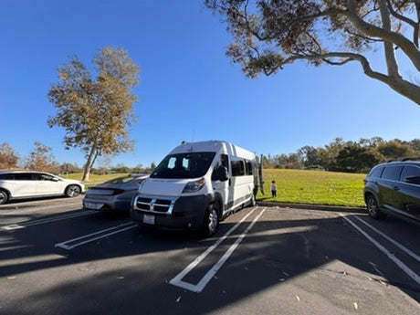White RV rental parked in a lot beside a grassy park with trees and a child, under a clear blue sky.
