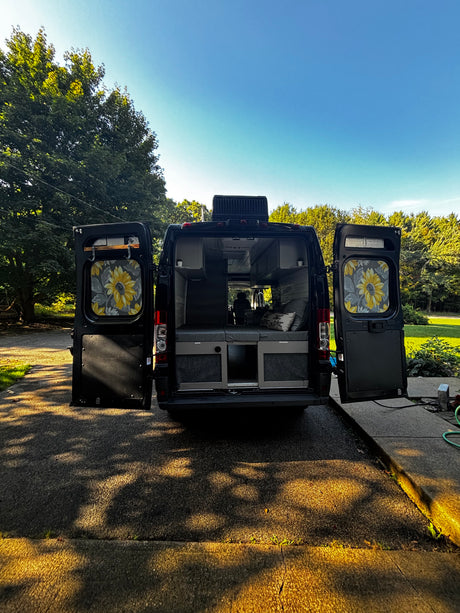 Open for adventure: a custom black camper van interior (potential rv rental) with unique sunflower window covers, parked on a sunny day.