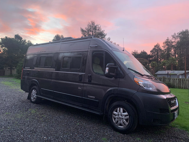 Dark gray Winnebago Travato camper van parked on gravel under a beautiful pink sunset. Ideal for your next rv rental.