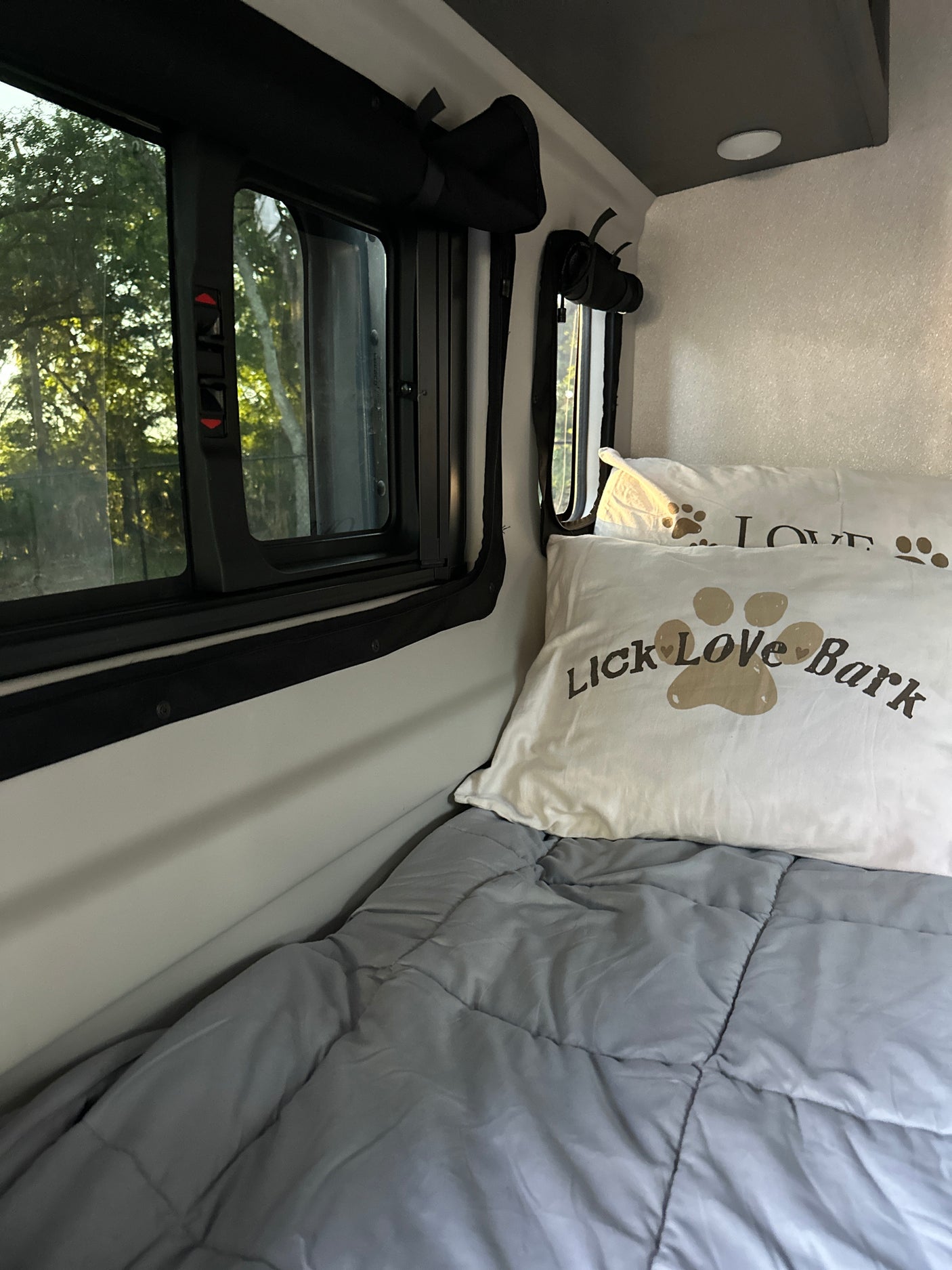 Cozy RV rental sleeping space with 'Lick Love Bark' pillows and a window view of lush trees outside.