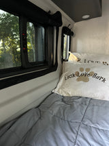 Cozy RV rental sleeping space with 'Lick Love Bark' pillows and a window view of lush trees outside.