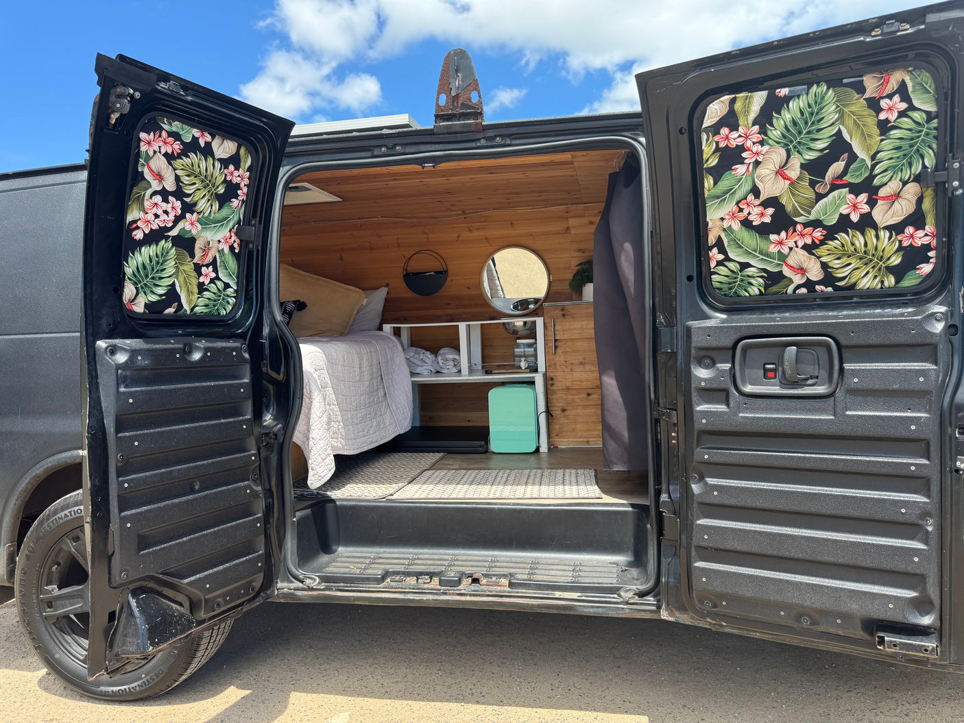 Inside an RV rental camper van. Open rear doors reveal a cozy bed, wood paneling, and tropical window coverings, ready for adventure.