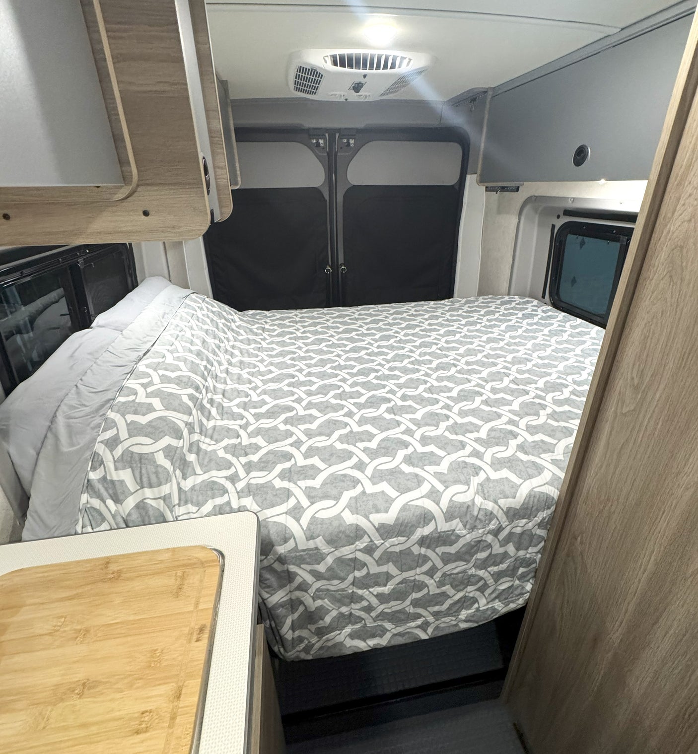 RV rental interior: cozy bed with grey pattern, ceiling AC unit, and light wood cabinetry.