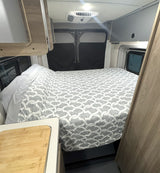 RV rental interior: cozy bed with grey pattern, ceiling AC unit, and light wood cabinetry.