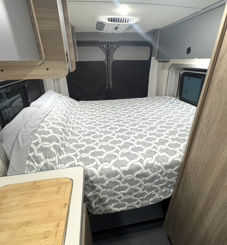 RV rental interior: cozy bed with grey pattern, ceiling AC unit, and light wood cabinetry.
