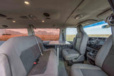 Inside an rv rental: grey passenger seats, a central table, and large windows revealing a scenic mountain-field landscape.