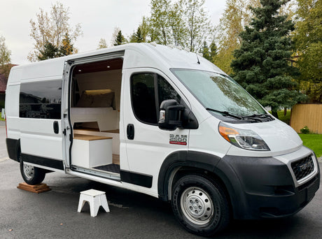 White Ram ProMaster camper van, ideal for rv rental. Open door reveals a cozy, custom-built interior with a bed & lighting.