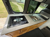 RV rental interior: A functional kitchen with Dometic stove, stainless steel sink, wood cabinets, and street view.