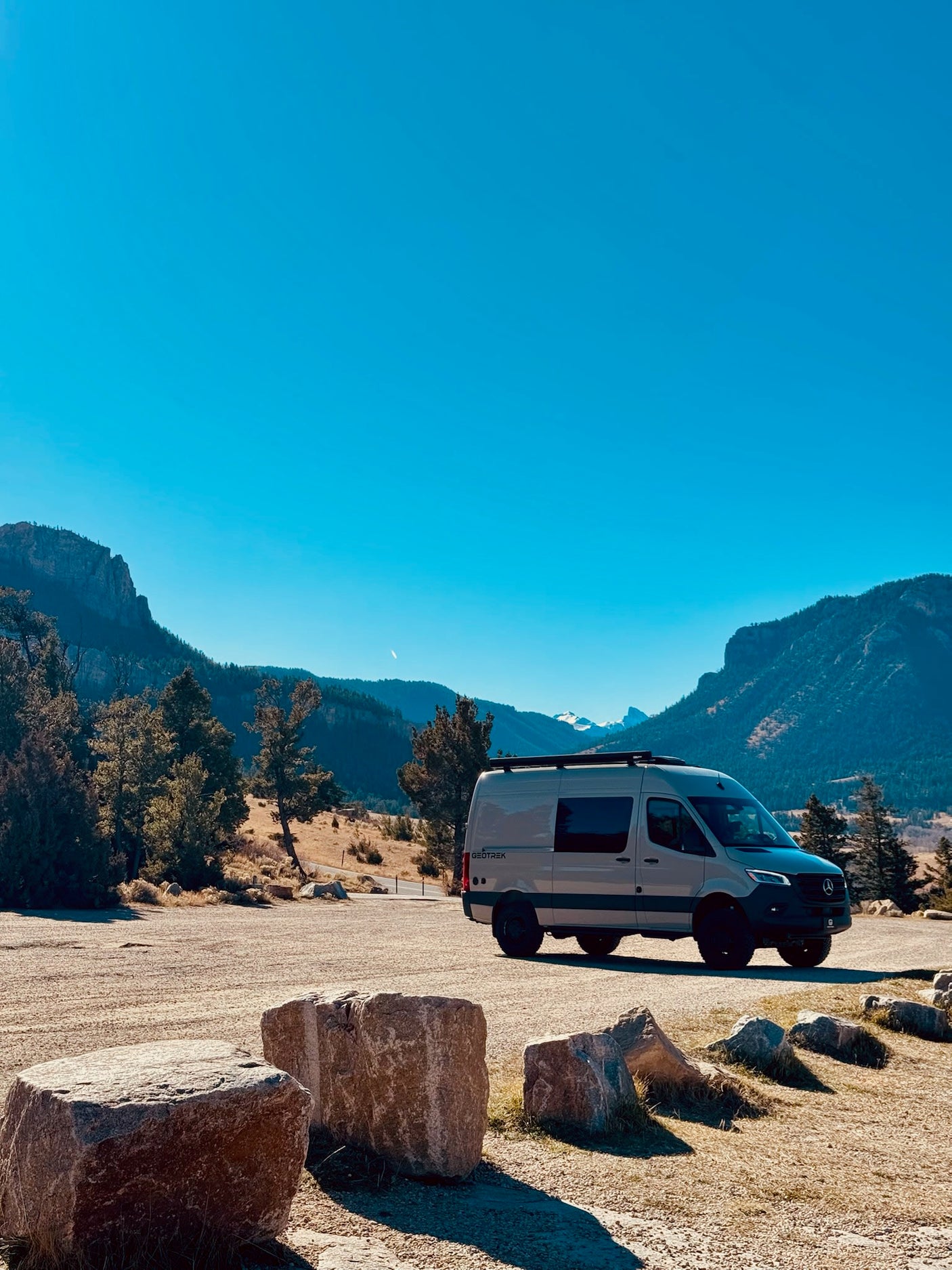 An rv rental camper van, branded Geotrek, parked in a scenic mountain landscape under a clear blue sky, with large rocks upfront.