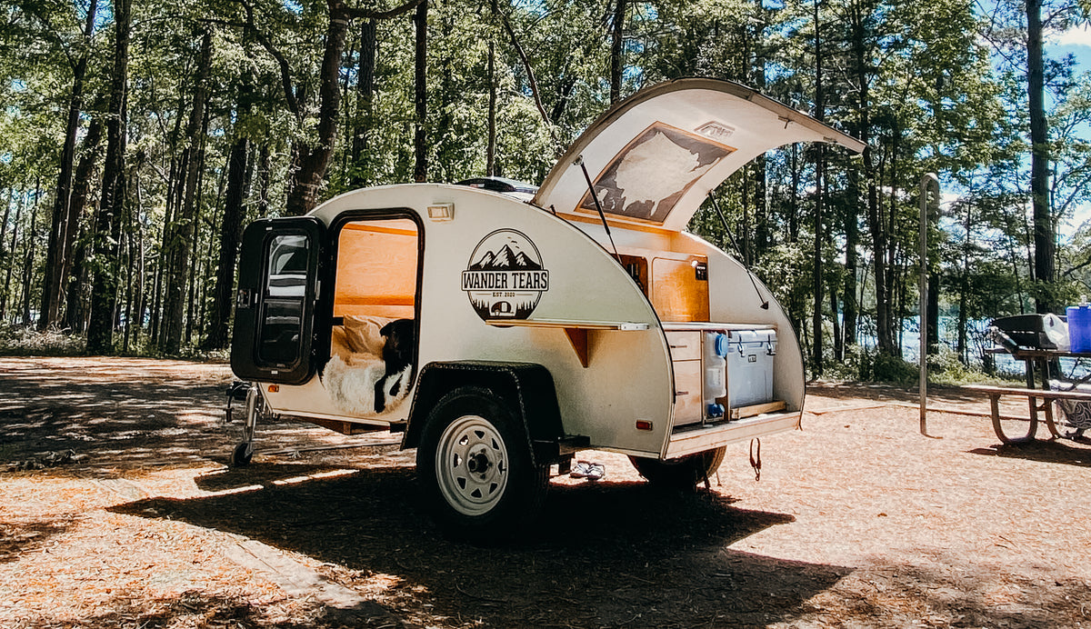 A compact white teardrop camper (an ideal RV rental) with its dog companion at a sunny forest campsite by a lake.