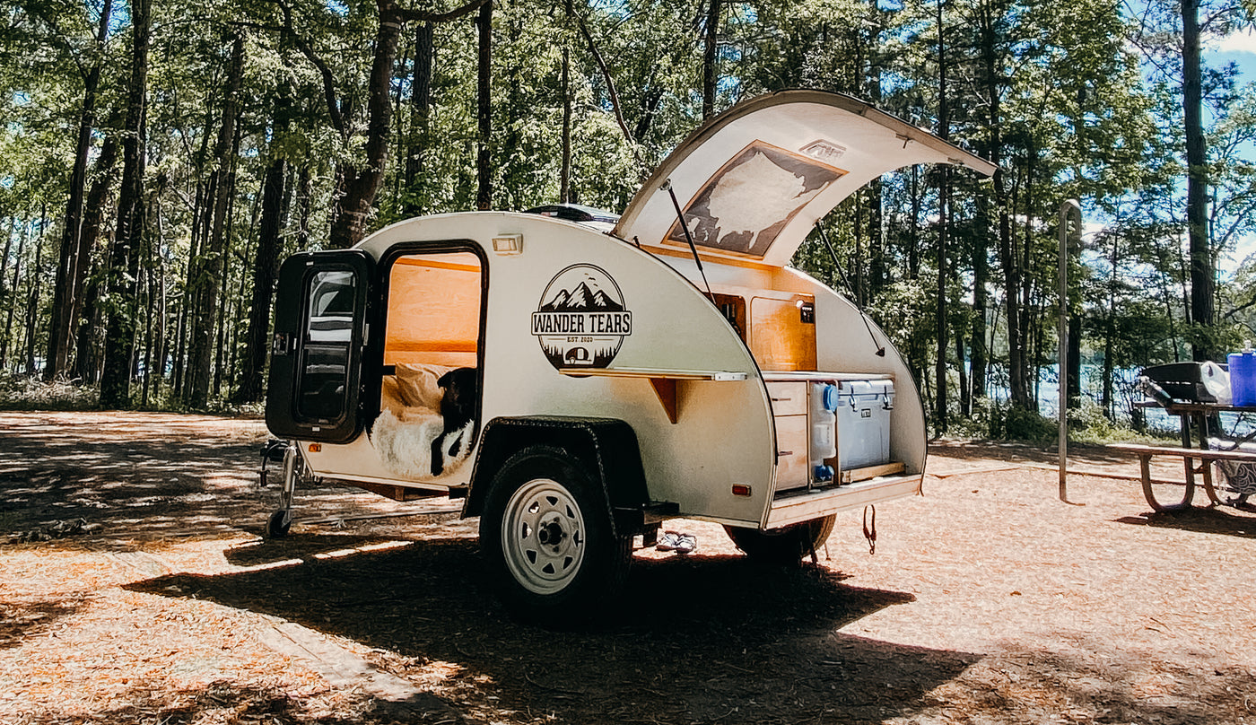 A compact white teardrop camper (an ideal RV rental) with its dog companion at a sunny forest campsite by a lake.