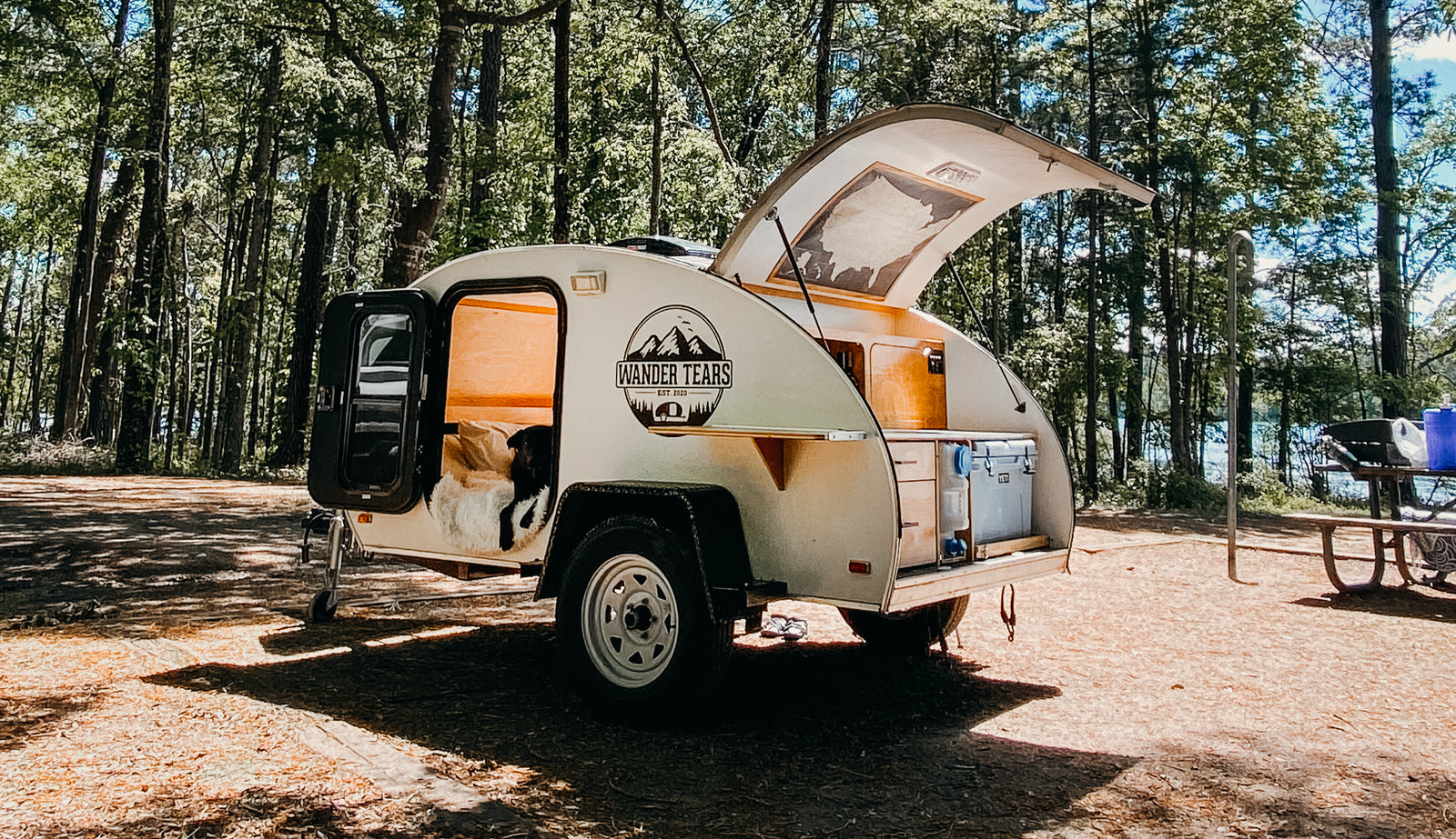 A compact white teardrop camper (an ideal RV rental) with its dog companion at a sunny forest campsite by a lake.