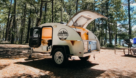 A compact white teardrop camper (an ideal RV rental) with its dog companion at a sunny forest campsite by a lake.
